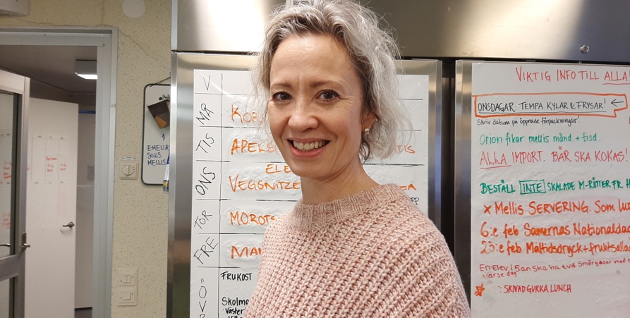 Kostchef Jenny Almroth står i köket på Volgsjö skola framför kylarna där veckans matsedel finns i bakgrunden. 