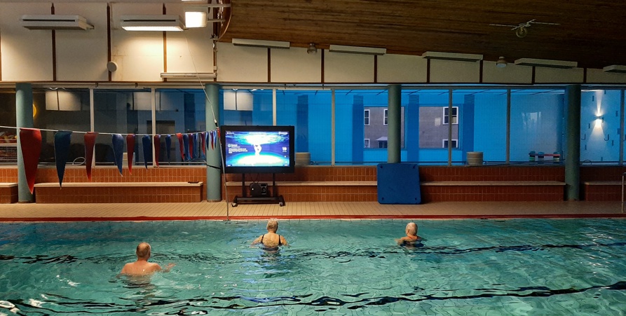 Tre deltagare i simbassängen som tränar vattengympa framför en tv-skärm.  