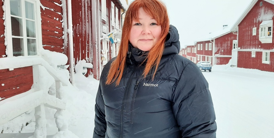 Foto på Carina Nybrand som står på Storgatan med de röda husen från Kyrkstan i bakgrunden. Carina har en blå vinterjacka och runt om är det mycket snö. 