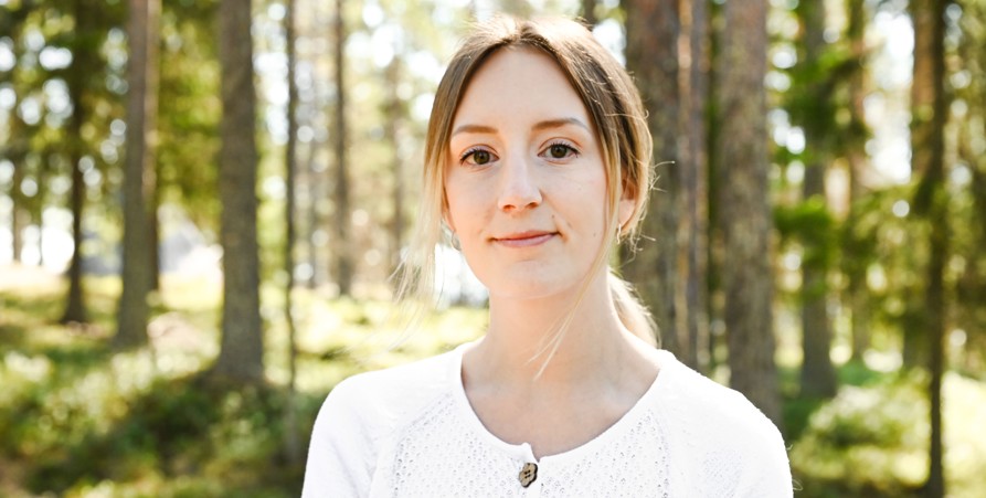 En ung kvinna med ljusbrunt hår, klädd i en vit tröja, står i ett soligt skogslandskap.