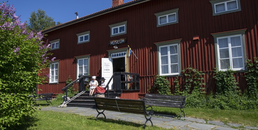 Foto på museet som är ett rött avlångt hus med vita fönster. På trappan in till museet sitter två personer. 