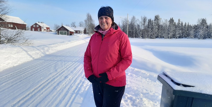 Anna Brunned står på en landsväg med röd vinterjacka på sig. I förgrunden syns ett sopkärl och i bakgrunden ett par röda hus. 